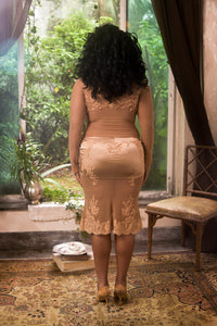 Woman in a beige dress standing in a room with a large window and plants.