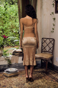 Woman in a beige lace dress standing in a room with a chair and plants.