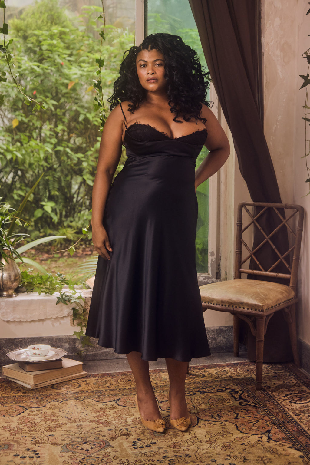 Woman wearing a black satin dress standing in a room with plants and a chair.