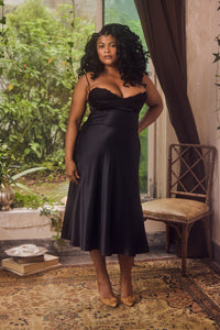 Woman wearing a black satin dress standing in a room with plants and a chair.