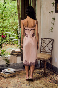Woman in a pink dress with lace details standing in a room with plants and a chair.